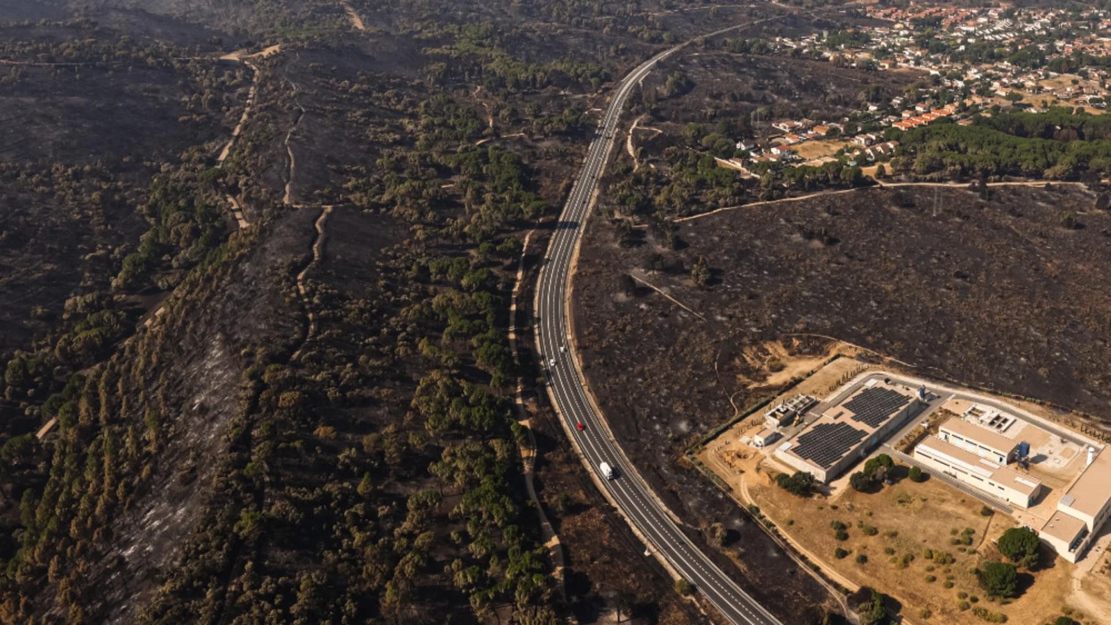 Vista aérea de una amplia zona quemada de la Sierra Oeste, atravesada por una carretera y junto a áreas urbanizadas y manchas de vegetación superviviente.