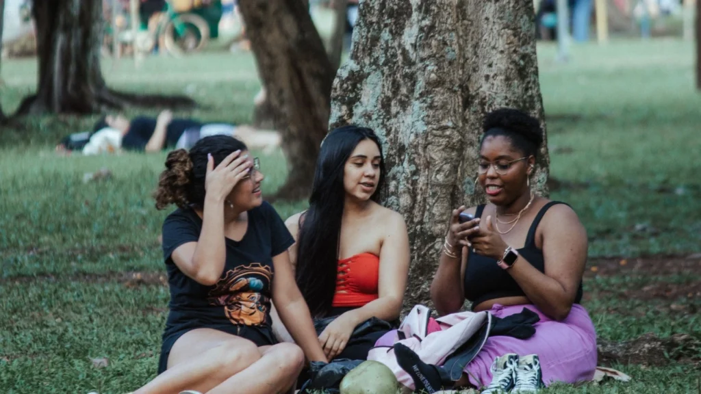 Tres mujeres sentadas sobre el césped bajo un árbol; una sostiene un teléfono móvil.