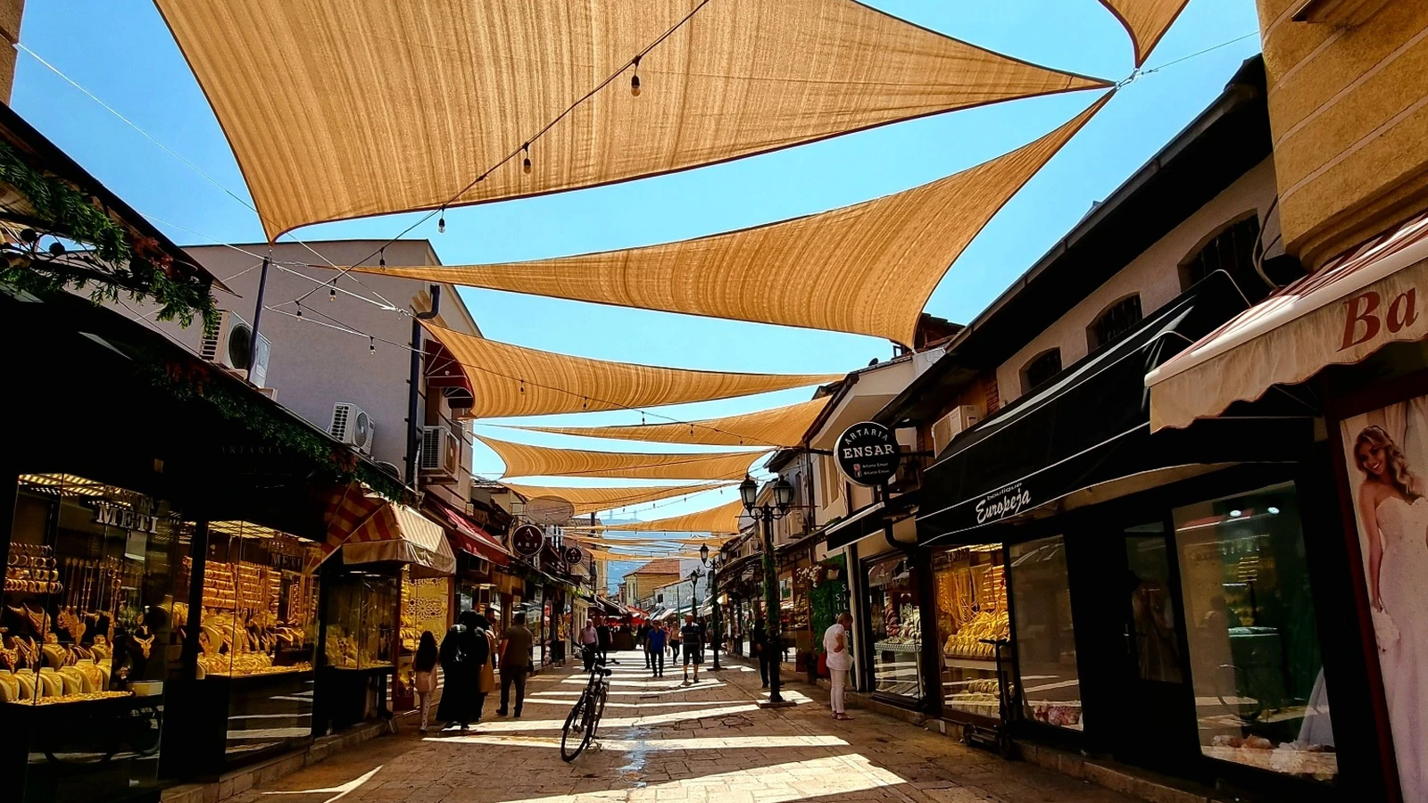 Toldos textiles triangulares dan sombra a una calle peatonal con comercios y viandantes.